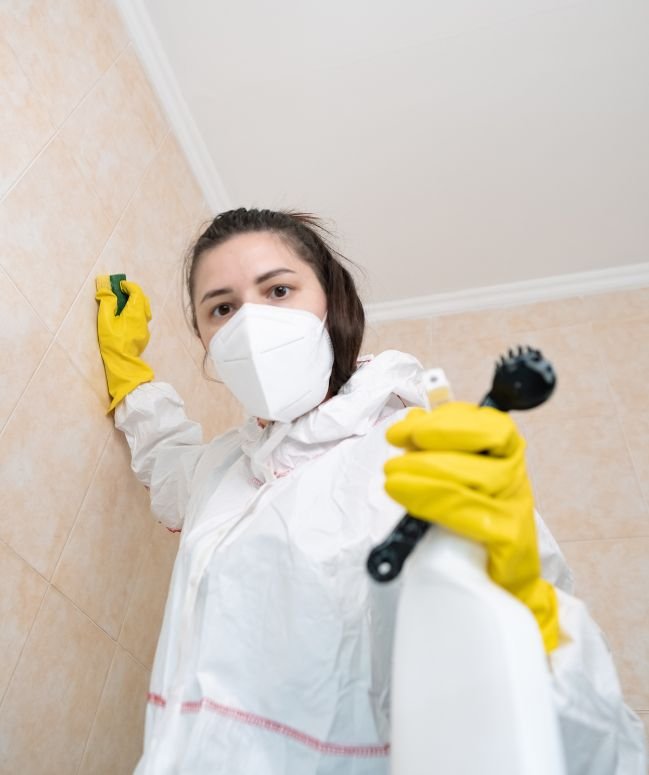 A woman in a white protective suit and yellow gloves holds a spray bottle for mold removal in Houston, TX.