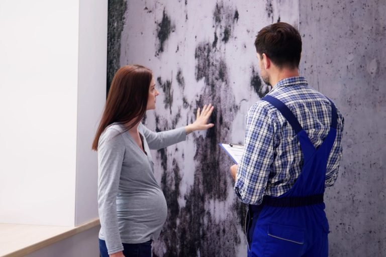 A man and woman stand together in front of a wall covered in mold, highlighting a neglected indoor environment.